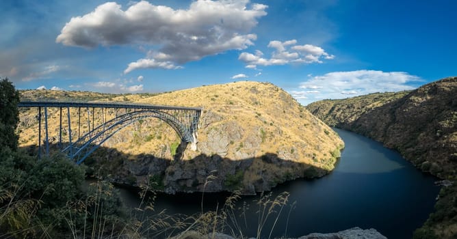 A scenic landscape with a steel bridge spanning a tranquil river, nestled among hills.
