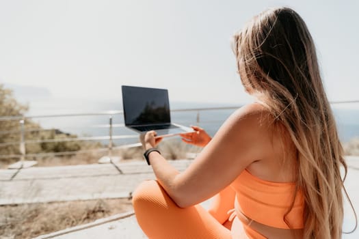 A woman in a bright orange sports bra and leggings uses a laptop while sitting on a balcony overlooking the sea.
