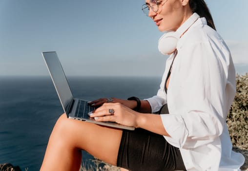 A woman wearing glasses and headphones sits on a cliff overlooking the ocean, typing on a laptop.