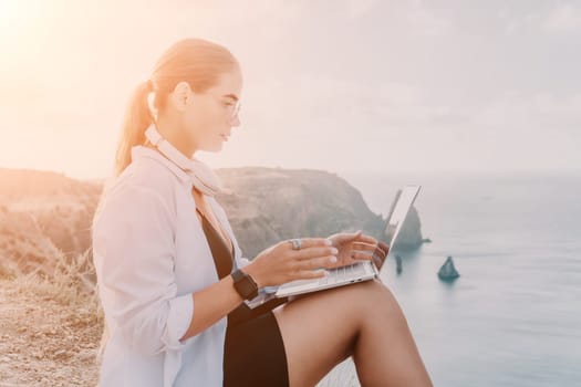 A woman sits on a cliff overlooking the ocean, working on a laptop. The sun shines brightly behind her.