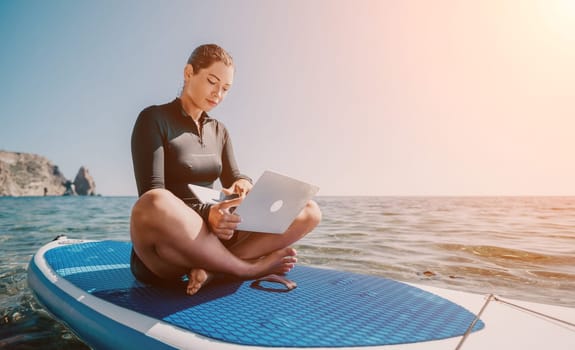 A woman sits in a lotus position on a paddleboard, working on a laptop. The water is calm and the sun is shining.