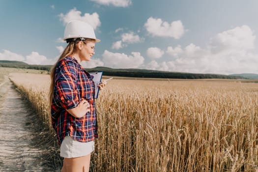 A young woman in a white hard hat and plaid shirt stands in a wheat field, examining the crops with a clipboard and pen.