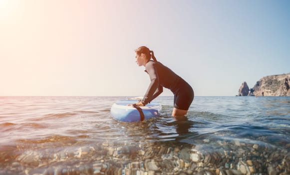 A woman in a wetsuit is preparing to paddle out on a surfboard in the ocean. The sun is shining brightly in the background.