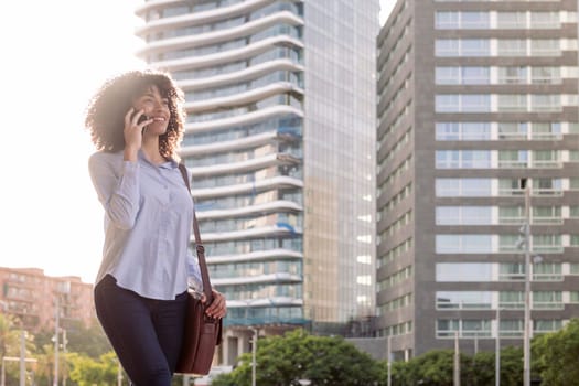 cheerful ethnic female entrepreneur standing in downtown and having conversation on mobile phone while looking away