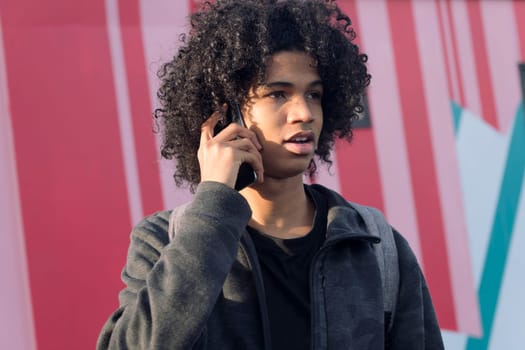 portrait of curly haired african american teenager talking on mobile phone in front of colorful wall, concept of technology of communication and young lifestyle