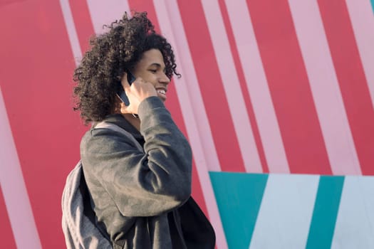 smiling curly haired african american teenager talking on mobile phone walking in front of colorful wall, concept of technology of communication and young lifestyle
