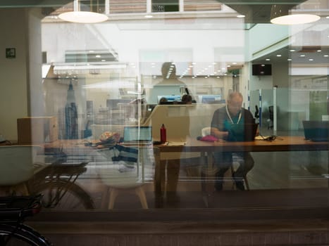 Cremona, Italy August 31st 2024 A senior manager is sitting at his desk in a modern open space office, working on his laptop, seen through the windows from the street