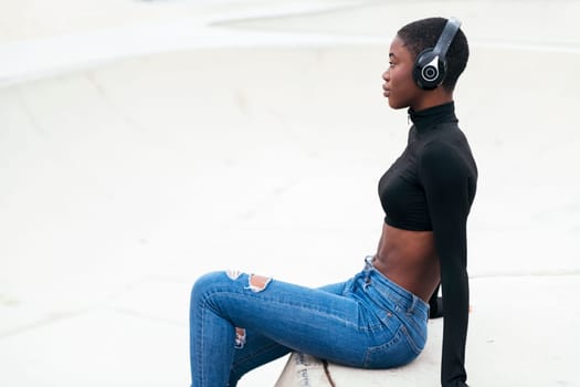 side view of young contemplative african american female in ripped jeans listening to music from wireless headphones while looking forward