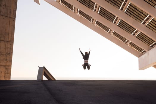 anonymous female athlete in sports clothes jumping with raised arms under modern construction with solar panels during workout, concept of urban sport and healthy lifestyle