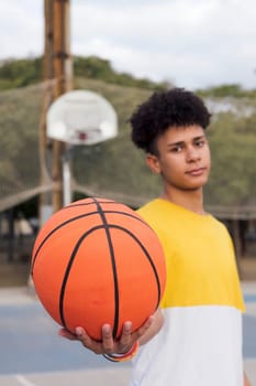 latin teen player showing basket ball standing on sports ground and looking at camera, concept of youth and urban lifestyle, selective focus on the ball