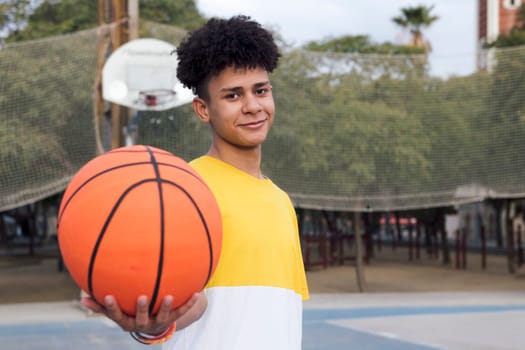 cheerful latin teen player showing basket ball standing on sports ground and looking at camera, concept of youth and urban lifestyle