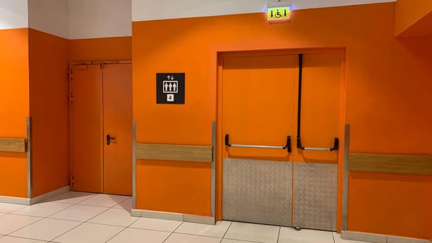 Corridor of building with orange walls next to door to elevator, a sign with admission of four people to occupancy of elevator and sign for disabled person for special elevator