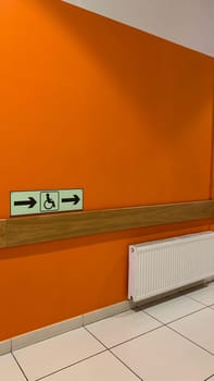 Vertical photo of orange wall in building with wheelchair sign with movement direction signs along corridor in access area for people with disabilities, way to wc with wheelchair access