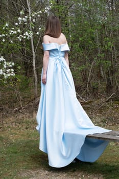 beautiful young woman in blue wedding dress against green forest background in early spring, standing on bench and showing long train of dress from behind, fashionable romantic style,