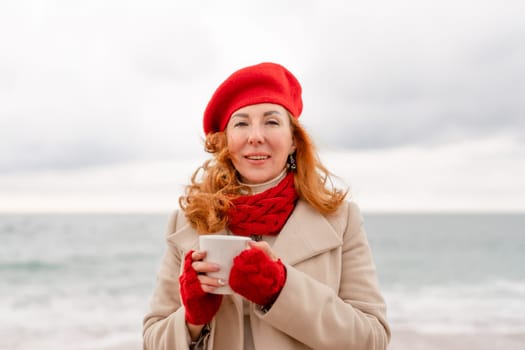 Woman beach in red beret scarf and mitts holding a cup of tea in his hands. Depicting beach relaxation and cozy attire. Walks by the sea.