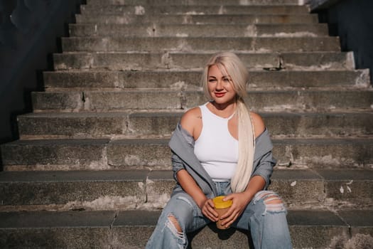 A blonde woman is sitting on a set of stairs, holding a cup of coffee. She is wearing a white tank top and jeans