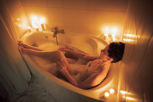A woman is sitting in a bathtub with candles lit around her. The candles are lit in a way that they create a warm and relaxing atmosphere
