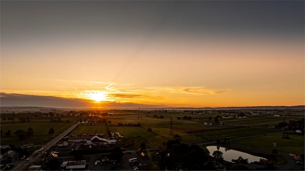 A beautiful sunset paints the sky with warm colors over a serene rural landscape. Fields stretch into the distance, while a calm pond reflects the fading light, creating a peaceful atmosphere.