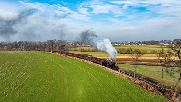 A charming steam train billows white smoke as it chugs along a picturesque landscape with vibrant green fields and a clear blue sky. . High quality photo