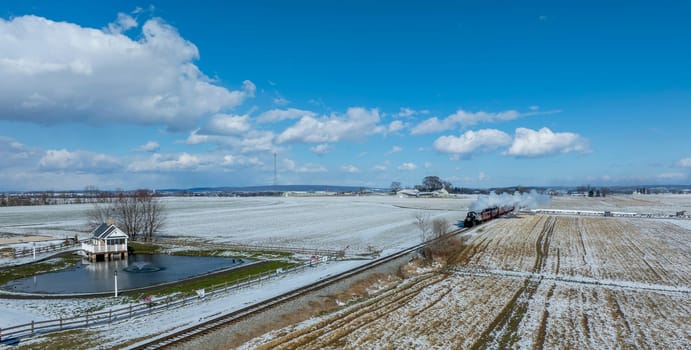 A steam locomotive chugs along the tracks, releasing vapor as it moves through snow-covered fields. In the foreground, a gazebo stands near a small pond, contributing to the serene winter landscape