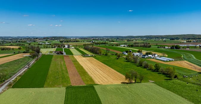Expansive farmlands display alternating fields of green and gold, with farms nestled among rolling hills under a bright blue sky, showcasing the beauty of rural agriculture.