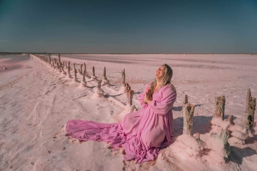 A woman in a pink dress sits on a wooden fence in a pink salt flat, gazing upwards at the sky.