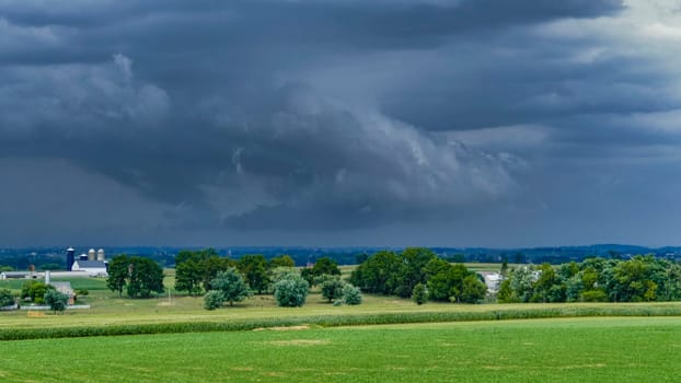 Threatening storm clouds gather above lush farmland, with a rustic barn and patches of trees visible. The atmosphere suggests an approaching storm in the rural landscape.