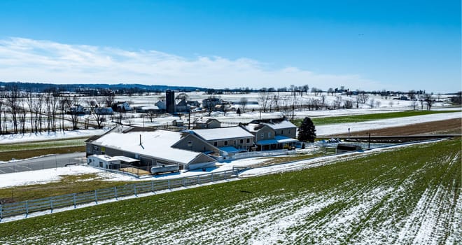 A picturesque rural farm features a snow-covered roof and green fields under bright blue skies, surrounded by snow-dusted landscapes and distant farms in the distance.