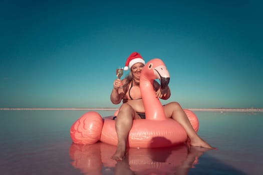A woman wearing a Santa hat sits on a pink flamingo float in a body of water. He is smiling. There are white posts in the background.