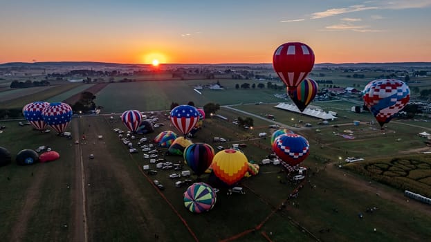 A dozen colorful hot air balloons are preparing for launch in a festival field during sunset. Spectators gather to witness the spectacle as the evening sky radiates warm colors.