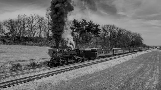 A classic steam train travels along tracks, billowing dark smoke against a backdrop of trees and fields. The scene captures the nostalgia of train travel amid nature's beauty, in Black and White