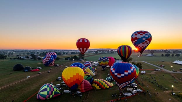 A dozen colorful hot air balloons are preparing for launch in a festival field during sunset. Spectators gather to witness the spectacle as the evening sky radiates warm colors.