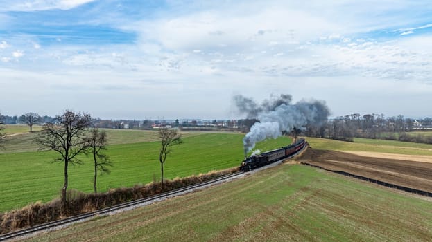 A steam locomotive chugs along train tracks surrounded by vast green fields and distant trees, emitting a plume of smoke against the cloudy sky, showcasing a tranquil rural setting.