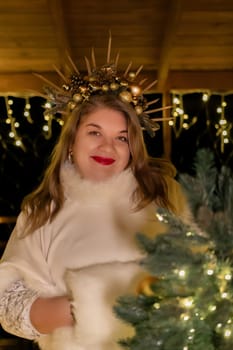 A woman with a golden crown stands in front of a Christmas tree, wearing a white sweater and fur shawl. She has long blonde hair and red lipstick. String lights illuminate the scene.
