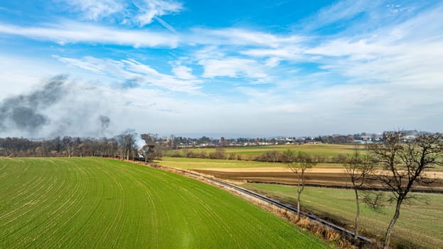Vibrant green fields stretch across the landscape, contrasting with patches of brown earth. In the distance, wisps of smoke rise against a clear blue sky, suggesting nearby agricultural activity.