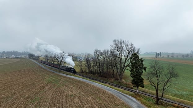 A vintage steam train glides along railway tracks bordered by fields and trees under a cloudy, fog-laden sky. The landscape showcases a serene rural environment in early morning light.