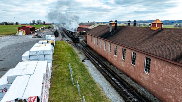 A vintage steam locomotive travels down the tracks adjacent to a historic brick station surrounded by green fields. The sky is overcast, creating a serene atmosphere in the area.
