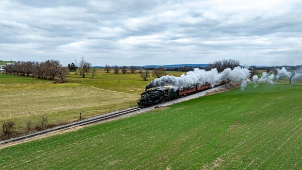 A classic steam train glides along the tracks, releasing plumes of white smoke while passing through lush green fields under a cloudy sky, highlighting the beauty of rural scenery. . High quality photo