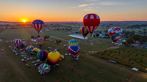 A dozen colorful hot air balloons are preparing for launch in a festival field during sunset. Spectators gather to witness the spectacle as the evening sky radiates warm colors.