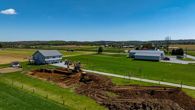Heavy machinery works on a construction site near a house, surrounded by fields under a clear blue sky. The area is bustling with activity, indicating progress on new development.