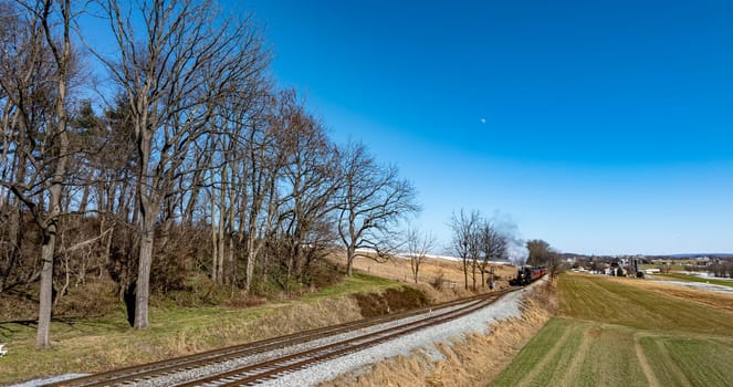 A vintage steam locomotive is making its way along the railway, surrounded by trees and rolling hills. The sky is clear and offers a stunning view of the scenery.