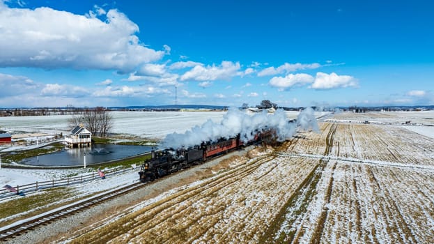 A steam train chugs through a snow-dusted landscape, offering a scenic winter journey by the rails, with a quaint pond house nearby.