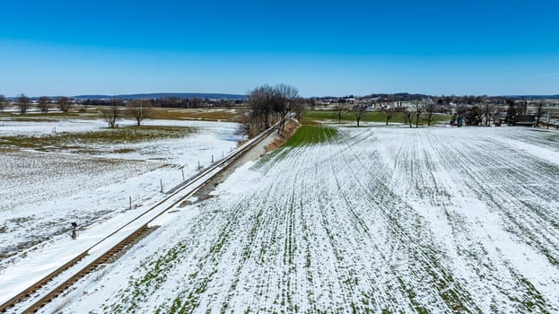 A vast expanse of snow-covered fields showcases the transition of seasons in a rural area, with hints of green emerging. The bright sky indicates a sunny, clear day, highlighting the serene landscape.