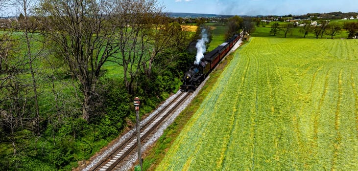 A steam locomotive chugs along railway tracks surrounded by vibrant green fields under a clear blue sky. The scene captures the charm of rural transportation in the countryside.
