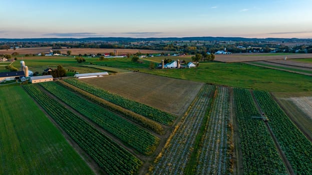 The sun sets over a picturesque rural landscape, highlighting expansive fields of crops and farmhouses scattered throughout. The vibrant green and brown layers form a patchwork effect.
