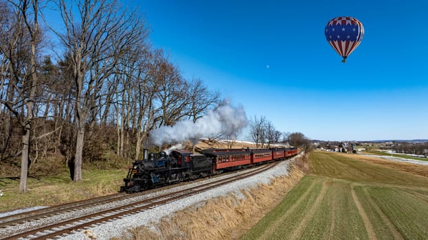 A vintage steam locomotive chugs along the tracks, releasing white smoke as it journeys through lush greenery