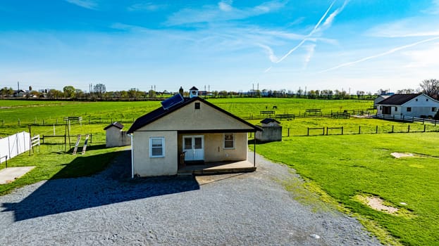 An Amish one room school house sits on a gravel area, surrounded by expansive green fields, barns, and distant structures under a bright blue sky.