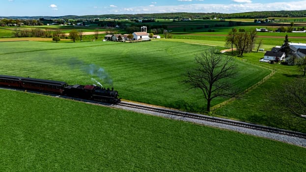 A vintage steam train chugs along a winding track, surrounded by vibrant green fields and rustic farmhouses. The sun shines brightly, highlighting the picturesque rural landscape.