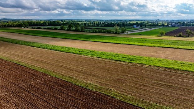 Expansive farmland stretches across the landscape, featuring alternating crop rows in varying shades of green and brown. Cloudy skies hover above the rich agricultural fields.