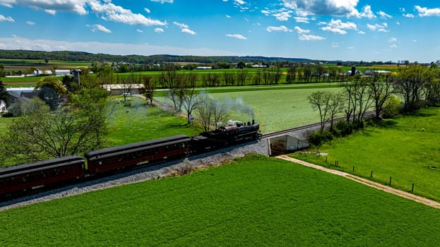 A steam locomotive moves along tracks bordered by vibrant green fields and trees under a clear blue sky. The scenic view captures the charm of countryside rail travel.